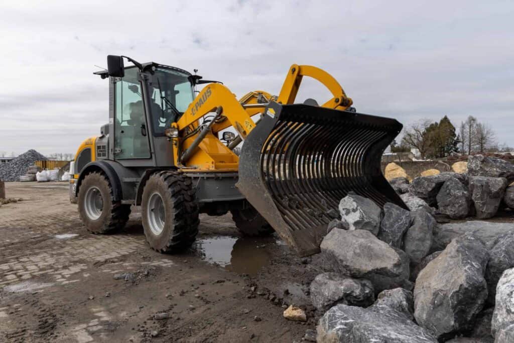 Yellow front-end loader with a large curved bucket at a rocky construction site, preparing to move boulders and gravel on a dirt surface.
