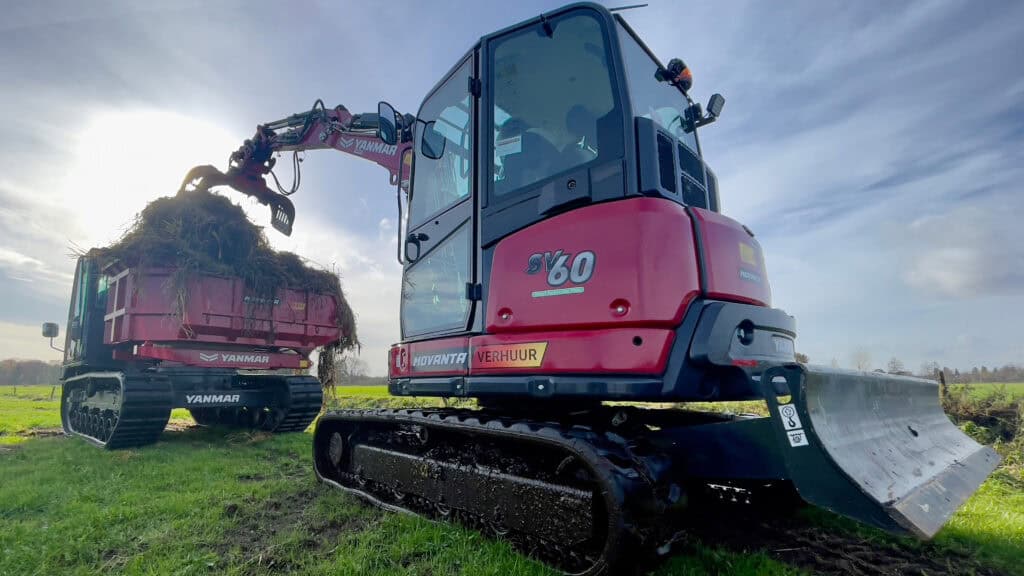 Kleine graafmachine op groene weide met zon en bewolkt hemel, geschikt voor bouw en tuinwerkzaamheden.