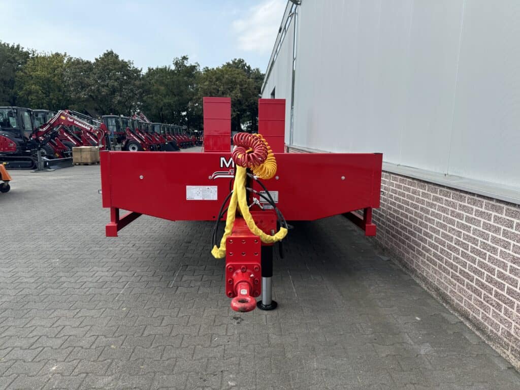 Robuuste rode oplegger voor bouwmachines met beveiligingskabels en toebehoren, opgesteld naast een industriële gevel.