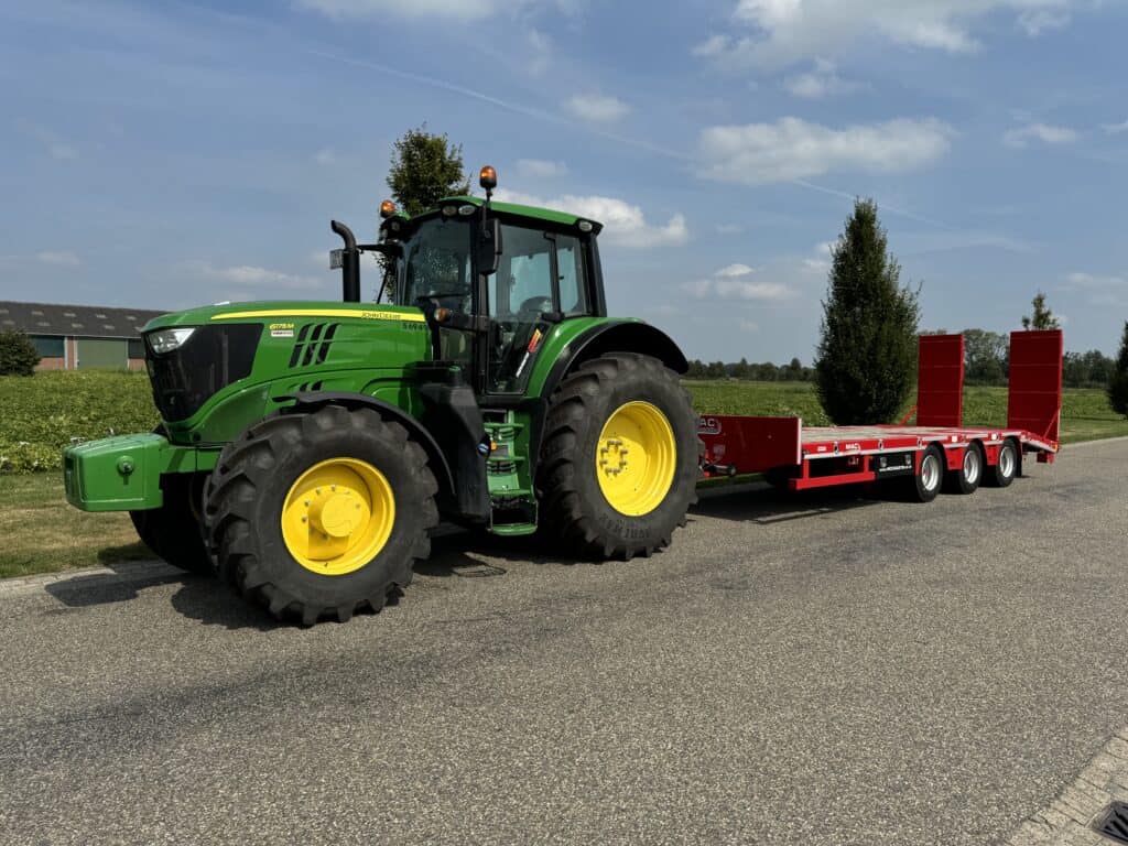 Groene John Deere tractor met rode transportplaten op een weg in een landelijke omgeving.