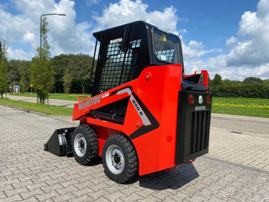 Manitou skid steer loader op parkeerterrein in zon, met groene omgeving op achtergrond.