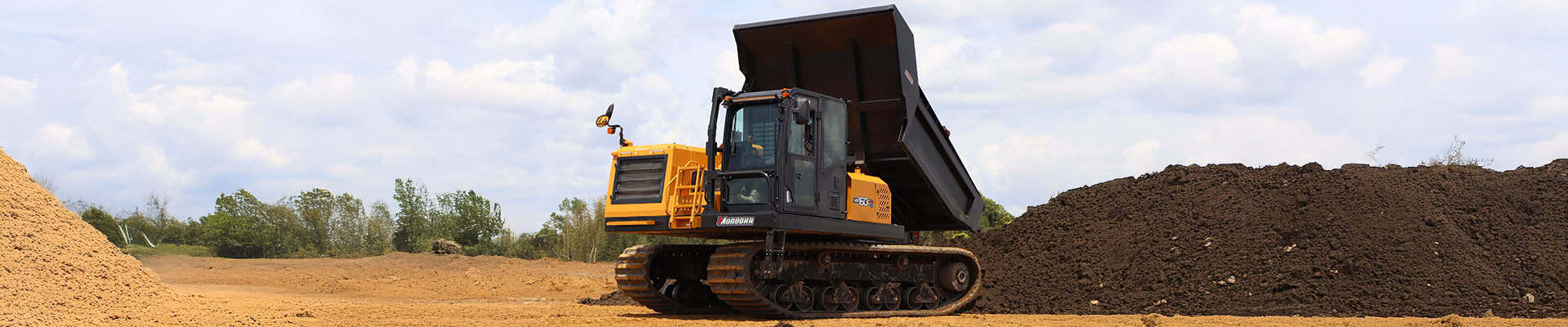 Large yellow tracked excavator with its dump bed raised, working on a dirt-site with mounds of soil nearby.