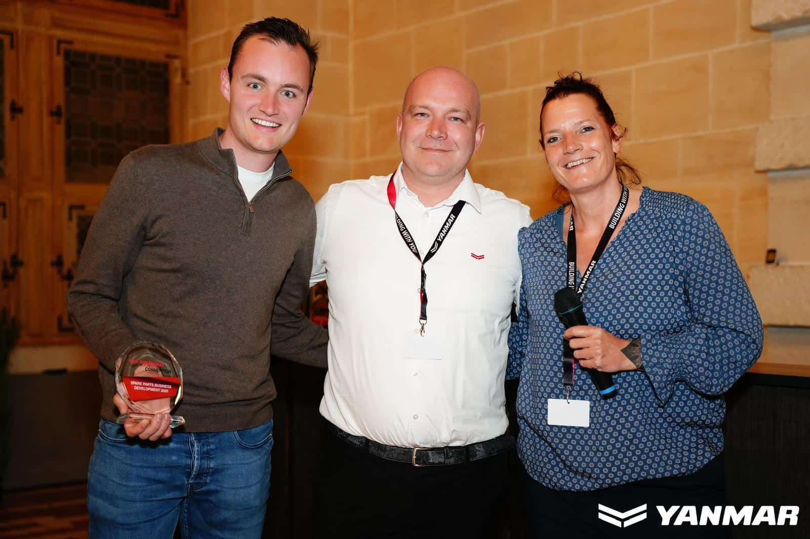 Three smiling colleagues posing for a photo at an awards event; the man on the left holds a glass trophy with a red plaque, flanked by a man in a white shirt and a woman with a microphone on the right.