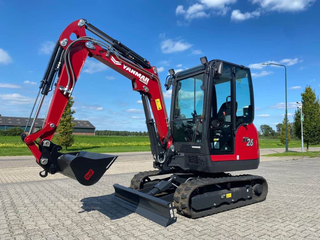 Kleine graafmachine Yanmar SV26 op een parkeerterrein met blauwe lucht en groene omgeving.