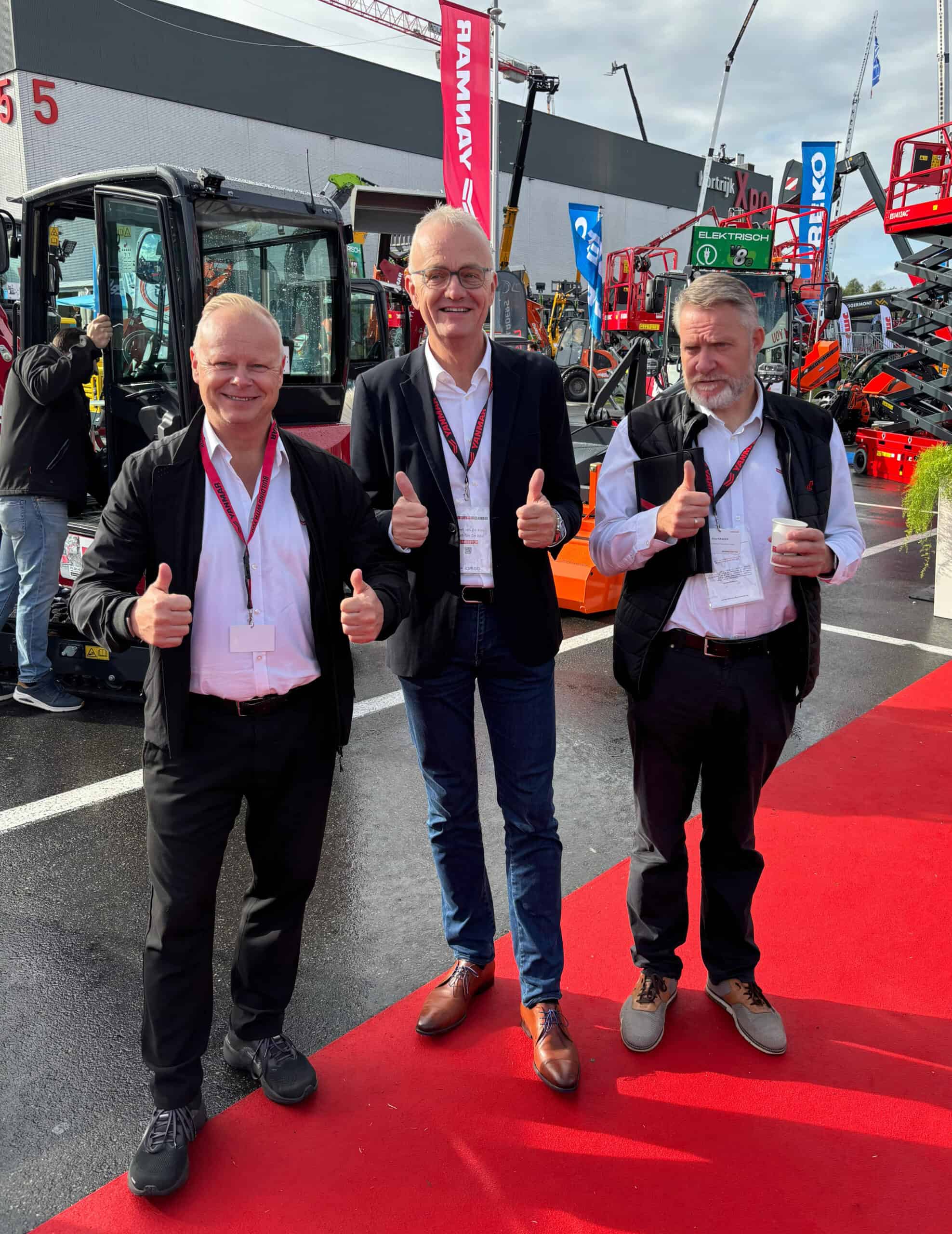 1. Drie mannen op bouwmachinebeurs met hand op schouder en duimen omhoog.